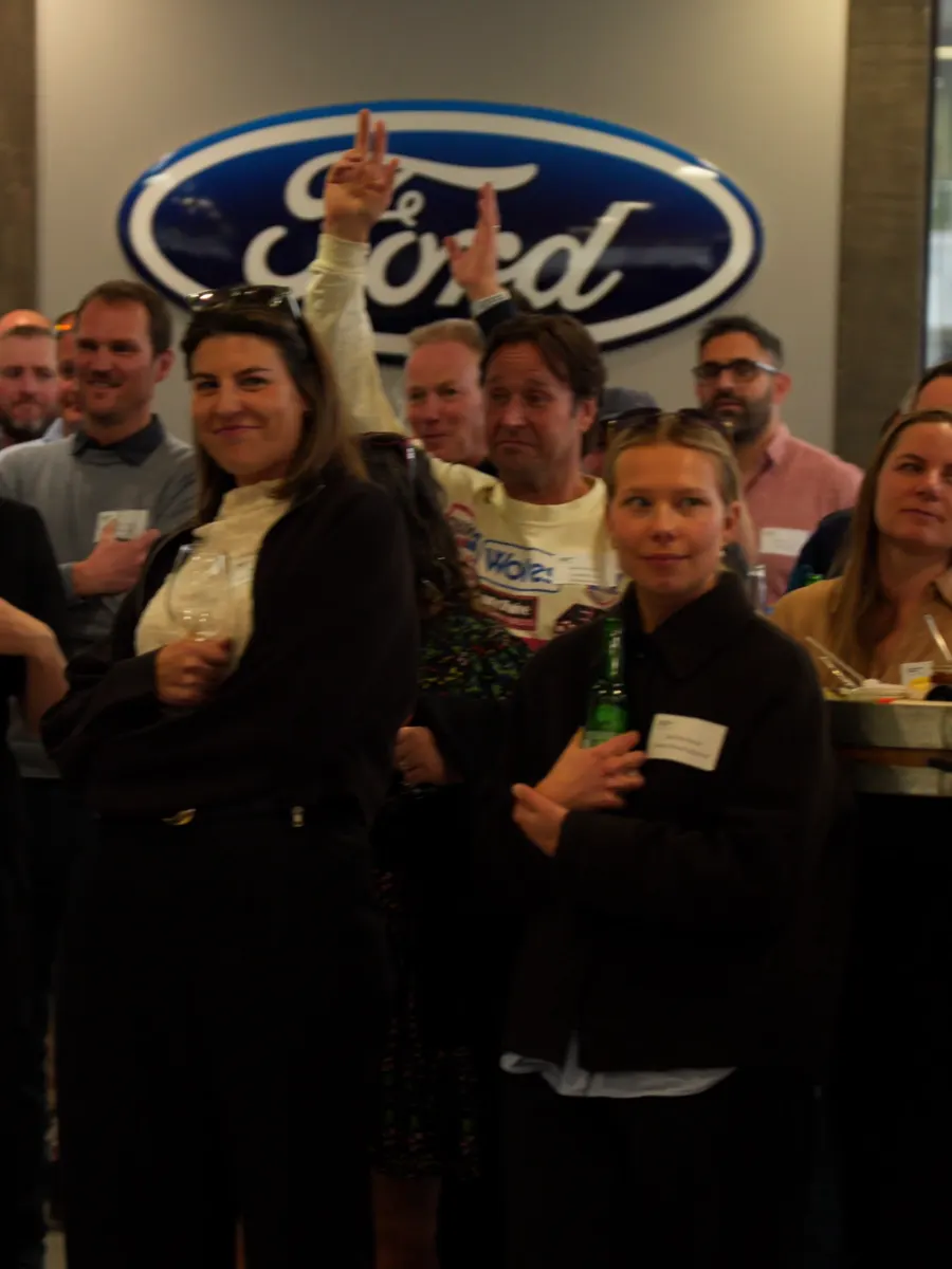 The crowd in the Macaulay Motors showroom laughing, one guest with a hand raised, under the Ford badge