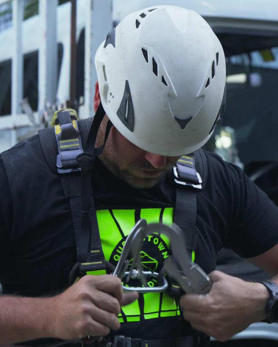A Queenstown Scaffolding crew member in a white helmet checking the clips on his harness before going up
