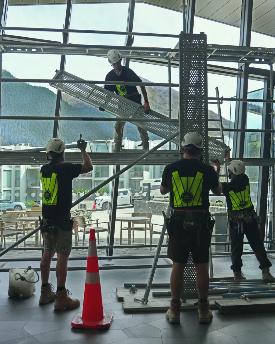 Three Queenstown Scaffolding crew in hi-vis building the scaffold inside the voco Queenstown lobby, with the mountains through the glass