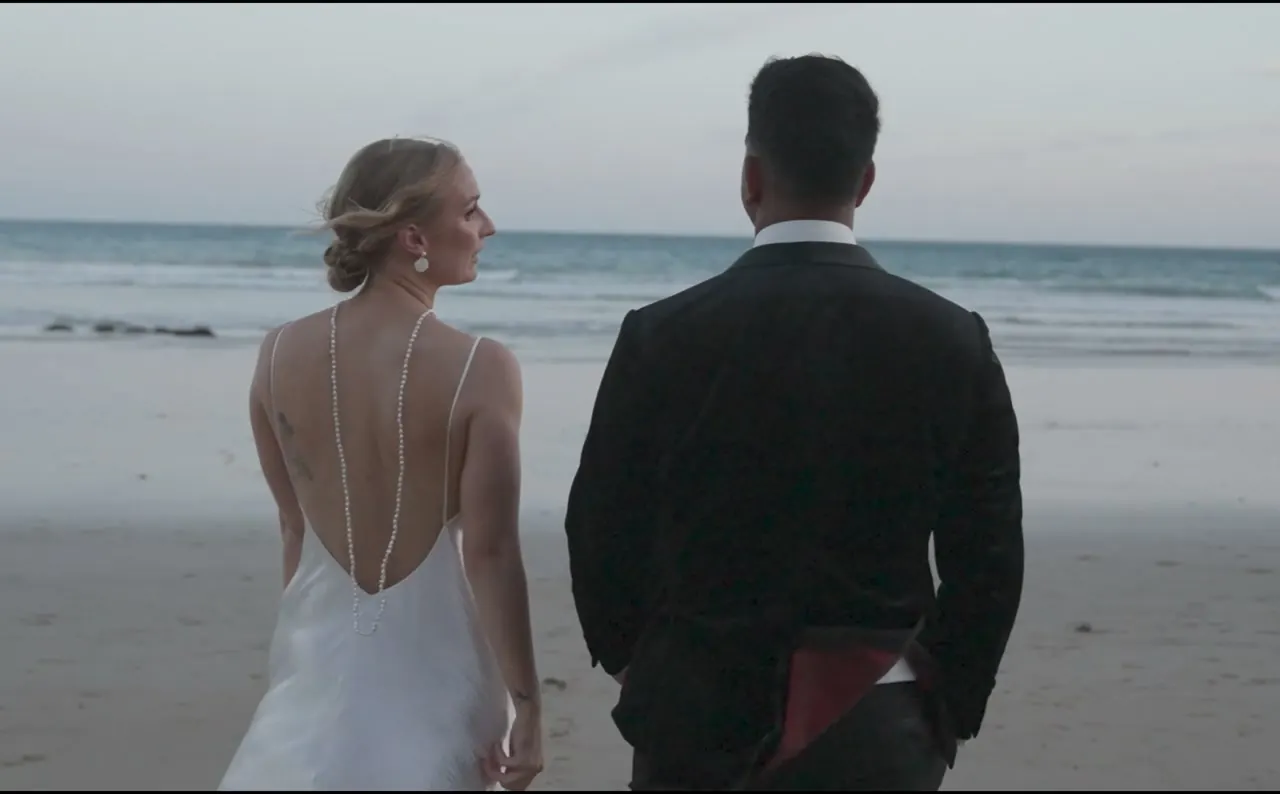 A bride and groom walking away from the camera along wet sand at dusk, the bride glancing back
